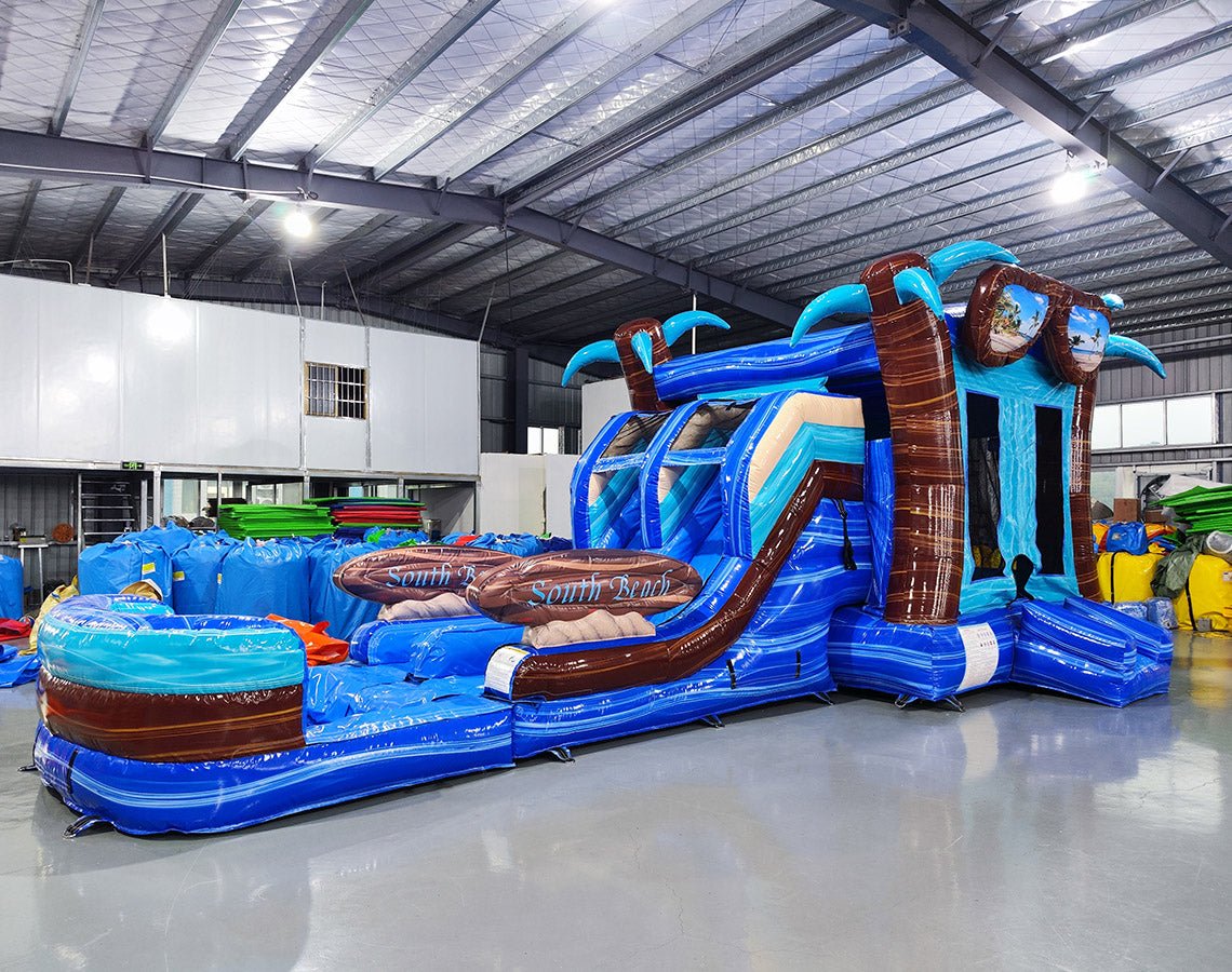 Three-quarter view of South Beach Dual Lane Commercial Inflatable Combo (Wet/Dry) set up in a warehouse. The inflatable features dual blue slides, brown palm tree pillars, a turquoise bounce house, and a blue splash pool base. Large sunglasses with beach scenes top the structure. Various other inflatable products and equipment are visible in the background, showcasing its commercial setting.