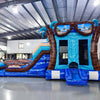 Side view of South Beach Dual Lane Commercial Inflatable Combo (Wet/Dry) showing the full length of the inflatable. Features dual blue slides, inflatable palm trees, a turquoise bounce house area, and a blue splash pool. Large inflatable sunglasses with beach scenes top the structure. "South Beach" is prominently displayed on the side. Set in a spacious warehouse with metal ceiling and concrete flooring.