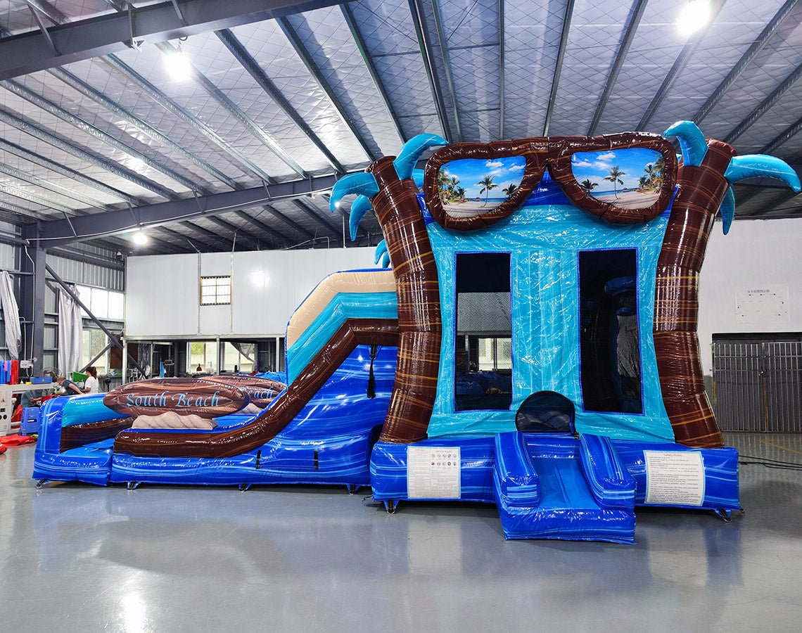 Side view of South Beach Dual Lane Commercial Inflatable Combo (Wet/Dry) showing the full length of the inflatable. Features dual blue slides, inflatable palm trees, a turquoise bounce house area, and a blue splash pool. Large inflatable sunglasses with beach scenes top the structure. "South Beach" is prominently displayed on the side. Set in a spacious warehouse with metal ceiling and concrete flooring.