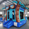 Angled view of the South Beach Econo Combo featuring a blue and turquoise bounce house with palm tree decorations, slide, and spacious splash pool. The inflatable includes a large sunglasses decoration on top and is set up in an indoor warehouse with other equipment and inflatables visible.