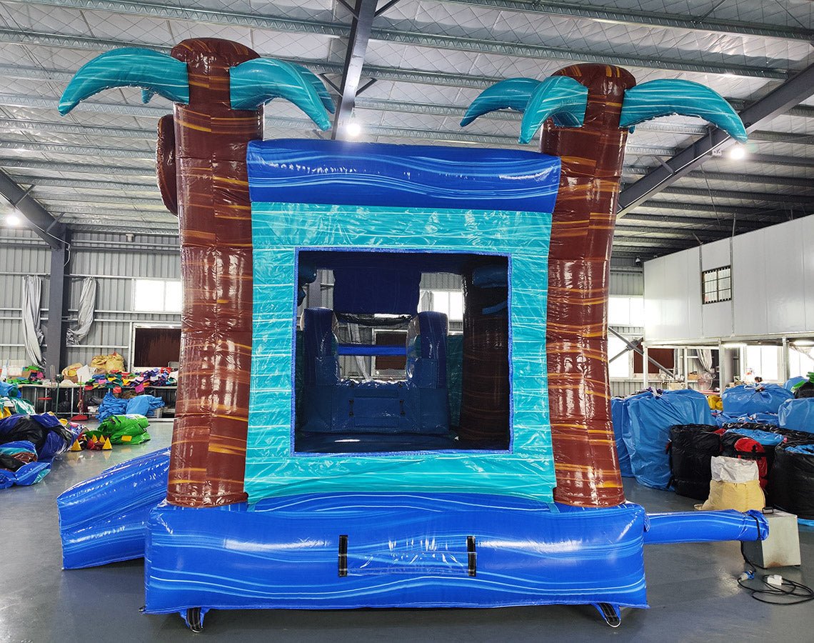 Front view of South Beach Econo Combo, showcasing the bounce house entrance flanked by inflatable palm trees, with a turquoise frame and blue base. Set up in an indoor warehouse with other inflatable products and equipment visible in the background.