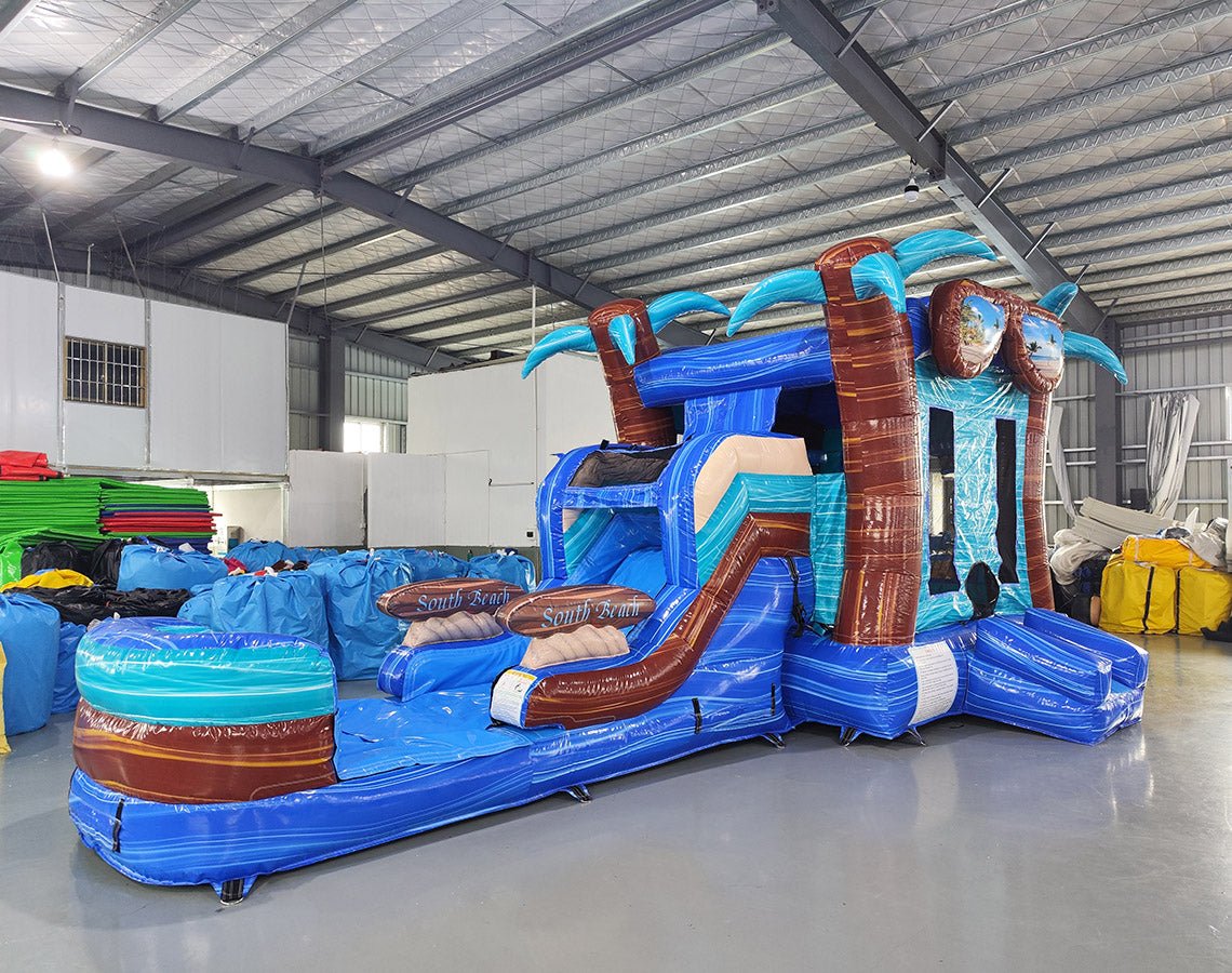 Large inflatable tropical-themed combo featuring a bounce house with palm tree decorations, attached slide, and splash pool. The structure has vibrant blue and turquoise colors with brown palm tree trunks and a large inflatable sunglasses decoration on top. Set up in an indoor warehouse with metal roofing and other inflatable products visible in the background.