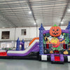 Side view of Spooky Splash Halloween Dual Lane Commercial Inflatable Combo featuring a haunted castle theme. The structure has blue castle towers, dual slides in red and purple, and a large orange jack-o'-lantern on top. A colorful Halloween scene with pumpkins and ghosts decorates the front panel. Set up in a large indoor warehouse with metal roofing and bright lighting visible.