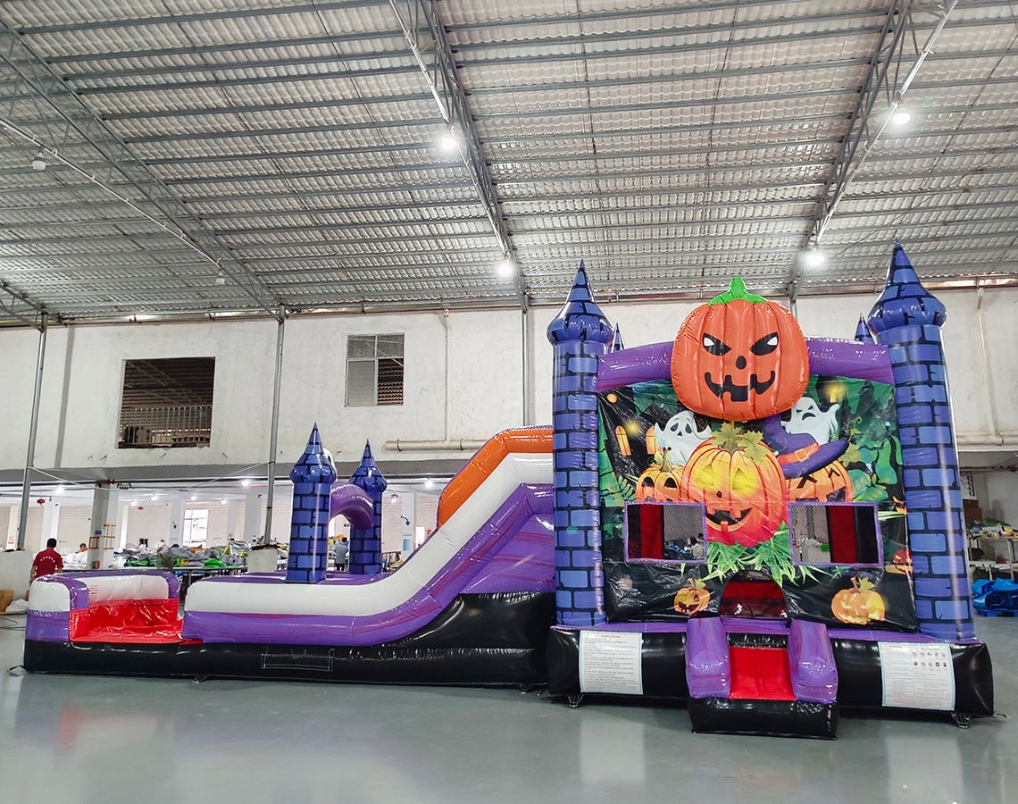 Side view of Spooky Splash Halloween Dual Lane Commercial Inflatable Combo featuring a haunted castle theme. The structure has blue castle towers, dual slides in red and purple, and a large orange jack-o'-lantern on top. A colorful Halloween scene with pumpkins and ghosts decorates the front panel. Set up in a large indoor warehouse with metal roofing and bright lighting visible.