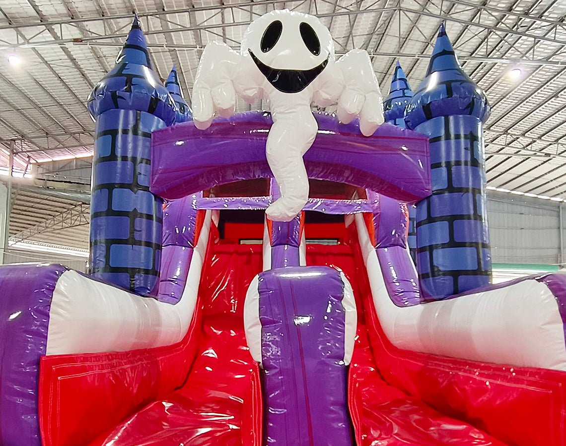 Close-up view of Spooky Splash Halloween Dual Lane Commercial Inflatable Combo showing the entrance area. Features blue castle towers, dual red slides, and a large inflatable ghost figure perched on top. The bounce house area has red entrances and purple walls. Set in a warehouse with metal roofing and bright overhead lighting.