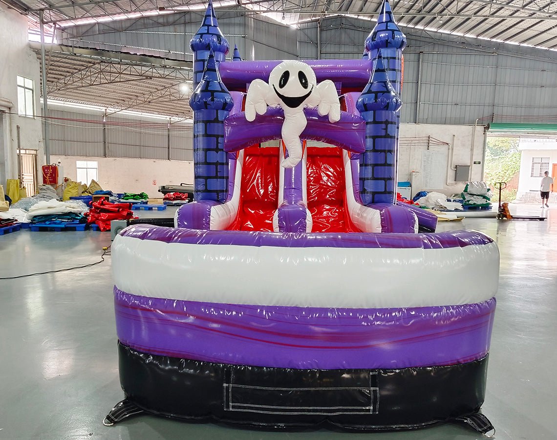 Wide-angle view of Spooky Splash Halloween Dual Lane Commercial Inflatable Combo (Wet/Dry) set up in a large indoor warehouse. The inflatable features blue castle towers, dual slides in red and purple, and a large orange jack-o'-lantern on top. A white inflatable ghost figure is perched between the slides. The front panel displays a colorful Halloween scene with pumpkins, ghosts, and other spooky elements. A white and purple splash pool area is visible at the base. The warehouse setting shows metal roofing,