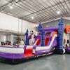 Side view of Spooky Splash Halloween Dual Lane Commercial Inflatable Combo set up in a large warehouse. The inflatable features blue castle towers, dual slides in red and purple, a large inflatable ghost, and an orange jack-o'-lantern on top. A white and purple splash pool area is visible at the base. Various other inflatable products and warehouse equipment can be seen in the background.