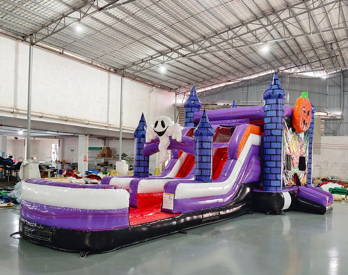 Side view of Spooky Splash Halloween Dual Lane Commercial Inflatable Combo set up in a large warehouse. The inflatable features blue castle towers, dual slides in red and purple, a large inflatable ghost, and an orange jack-o'-lantern on top. A white and purple splash pool area is visible at the base. Various other inflatable products and warehouse equipment can be seen in the background.