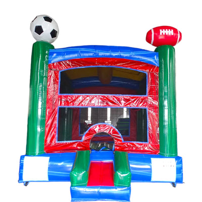 Sports-themed inflatable bounce house featuring green pillars with a soccer ball and football on top. The structure has a red front wall with mesh windows, blue side walls, and a blue roof. A small slide protrudes from the entrance. Safety instructions are visible on the base. The bounce house is set against a white background.