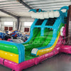 Three-quarter view of Summer Luau Dual Lane Commercial Inflatable Combo in a spacious warehouse. The inflatable displays dual slides in blue and green, inflatable palm trees, a turquoise bounce house structure with colorful flowers, and a rainbow-colored splash pool. Various other inflatable products and warehouse equipment can be seen, demonstrating its size and commercial application.