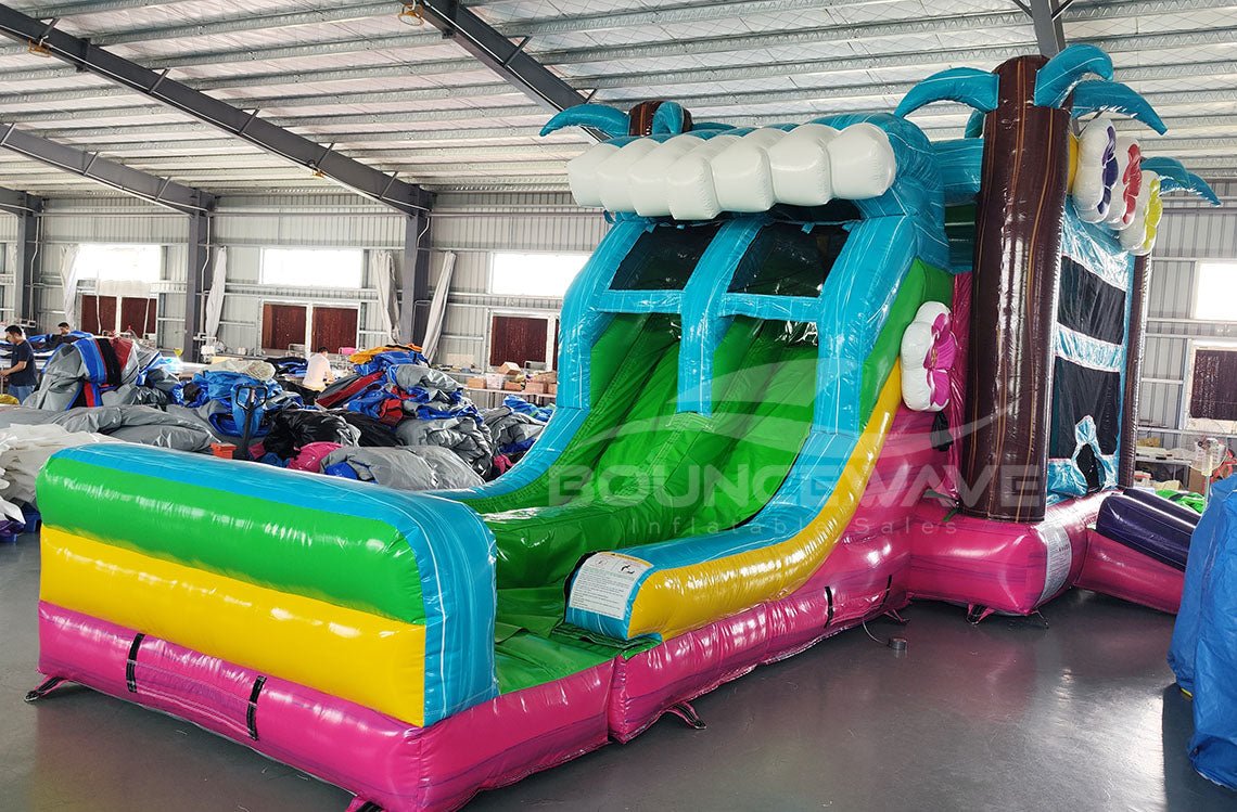 Three-quarter view of Summer Luau Dual Lane Commercial Inflatable Combo in a spacious warehouse. The inflatable displays dual slides in blue and green, inflatable palm trees, a turquoise bounce house structure with colorful flowers, and a rainbow-colored splash pool. Various other inflatable products and warehouse equipment can be seen, demonstrating its size and commercial application.