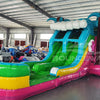 Wide-angle view of Summer Luau Dual Lane Commercial Inflatable Combo set up in a large indoor warehouse. The inflatable features dual slides in blue and green, inflatable palm trees, a turquoise bounce house area with colorful flowers, and a rainbow-colored splash pool. Metal roofing, support beams, and various other inflatable products are visible in the background.