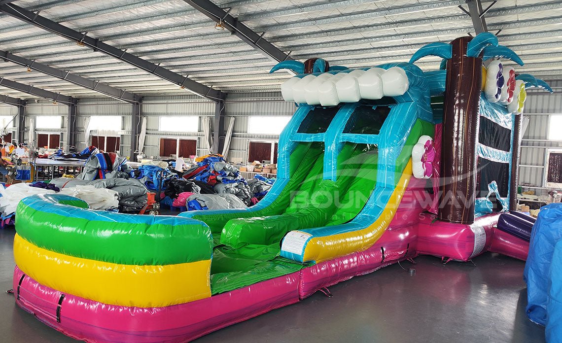 Wide-angle view of Summer Luau Dual Lane Commercial Inflatable Combo set up in a large indoor warehouse. The inflatable features dual slides in blue and green, inflatable palm trees, a turquoise bounce house area with colorful flowers, and a rainbow-colored splash pool. Metal roofing, support beams, and various other inflatable products are visible in the background.