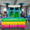 Front view of Summer Luau Dual Lane Commercial Inflatable Combo (Wet/Dry) set up in a large indoor warehouse. The inflatable features dual slides in blue and green, brown inflatable palm tree trunks with blue leaves, and a turquoise bounce house area decorated with colorful flower ornaments. The base has a vibrant rainbow color scheme of yellow, green, and pink. The "BOUNCEWAVE" logo is visible on the front. Various other inflatable products and equipment are visible in the background.