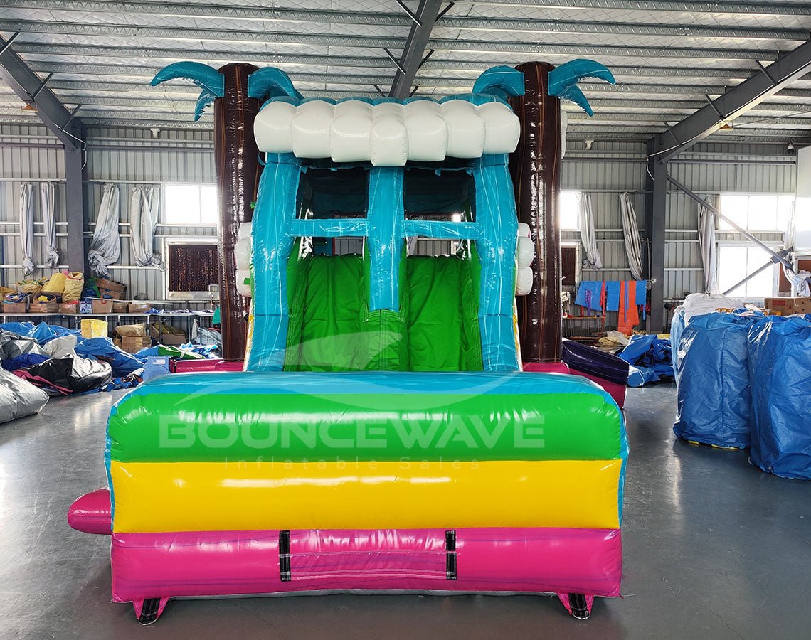 Front view of Summer Luau Dual Lane Commercial Inflatable Combo (Wet/Dry) set up in a large indoor warehouse. The inflatable features dual slides in blue and green, brown inflatable palm tree trunks with blue leaves, and a turquoise bounce house area decorated with colorful flower ornaments. The base has a vibrant rainbow color scheme of yellow, green, and pink. The "BOUNCEWAVE" logo is visible on the front. Various other inflatable products and equipment are visible in the background.