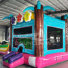 Cross Rear view of Summer Luau Dual Lane Commercial Inflatable Combo showing the bounce house entrance. Features a turquoise frame with mesh windows, brown inflatable palm tree trunks, and colorful flower decorations on top. The dual slides and splash pool are partially visible. Set in a warehouse with concrete flooring and various other inflatable products and equipment visible in the background.