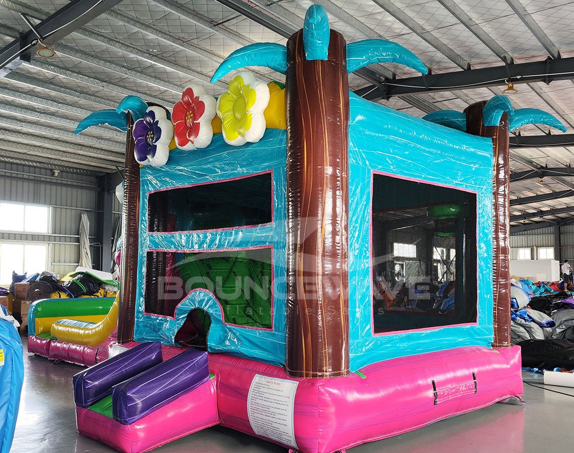 Cross Rear view of Summer Luau Dual Lane Commercial Inflatable Combo showing the bounce house entrance. Features a turquoise frame with mesh windows, brown inflatable palm tree trunks, and colorful flower decorations on top. The dual slides and splash pool are partially visible. Set in a warehouse with concrete flooring and various other inflatable products and equipment visible in the background.