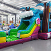 Wide-angle view of Summer Luau Econo Combo featuring a colorful bounce house with palm tree decorations, curved slide, and spacious splash pool. The inflatable has vibrant blue, green, yellow, and pink colors and is set up in an indoor warehouse with other equipment and inflatables visible.