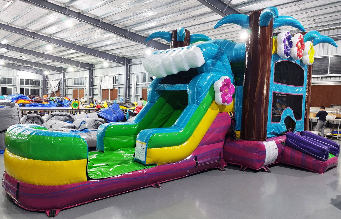 Wide-angle view of Summer Luau Econo Combo featuring a colorful bounce house with palm tree decorations, curved slide, and spacious splash pool. The inflatable has vibrant blue, green, yellow, and pink colors and is set up in an indoor warehouse with other equipment and inflatables visible.