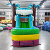 Large inflatable tropical-themed combo featuring a bounce house with palm tree decorations, attached slide, and splash pool. The structure has vibrant blue, green, yellow, and pink colors with brown palm tree trunks. Set up in an indoor warehouse with metal roofing visible.