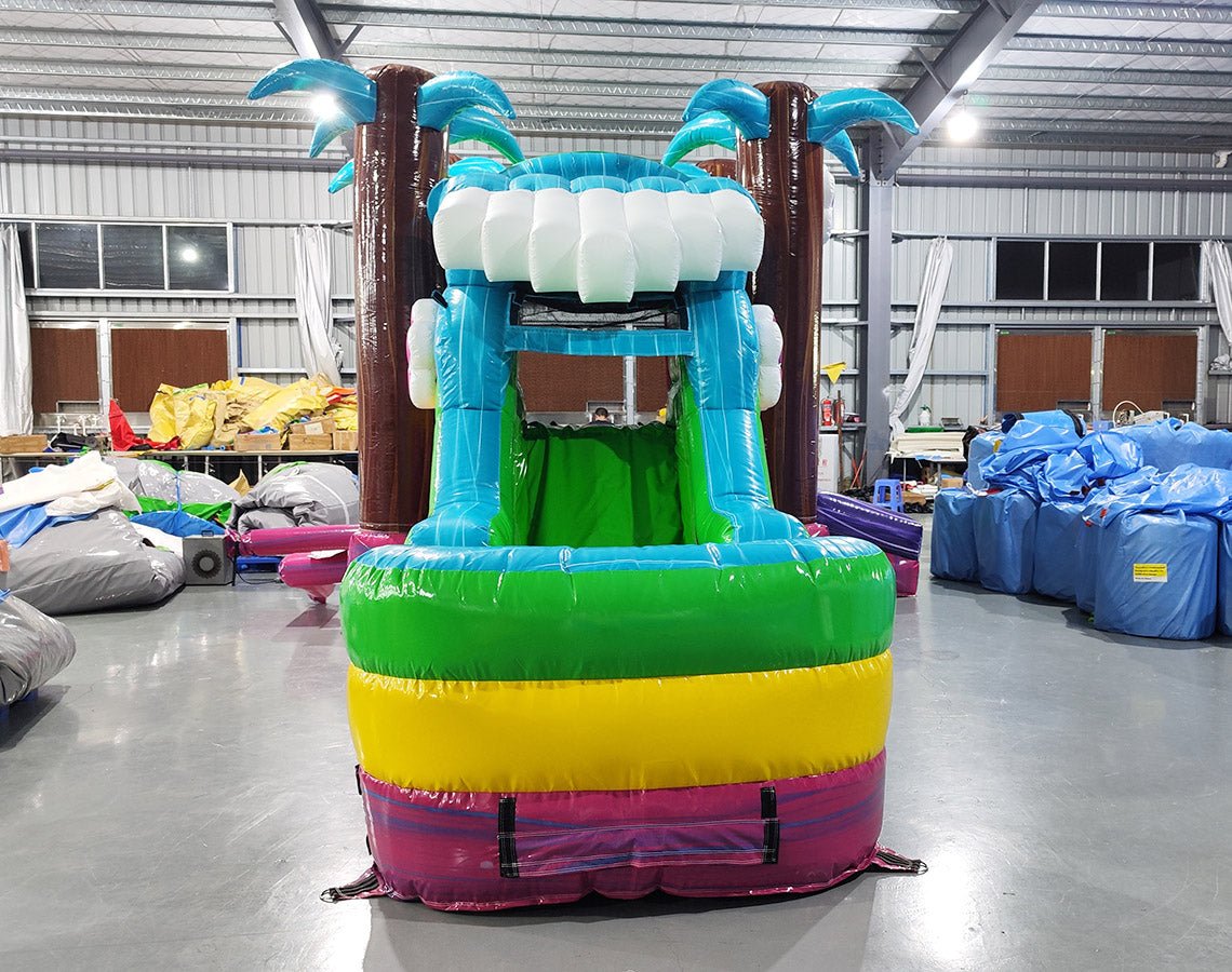 Large inflatable tropical-themed combo featuring a bounce house with palm tree decorations, attached slide, and splash pool. The structure has vibrant blue, green, yellow, and pink colors with brown palm tree trunks. Set up in an indoor warehouse with metal roofing visible.