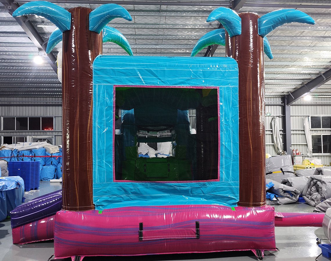 Front view of Summer Luau Econo Combo, showcasing the bounce house entrance flanked by inflatable palm trees, with a blue frame and colorful base. Set up in an indoor warehouse with other inflatable products and equipment visible in the background.