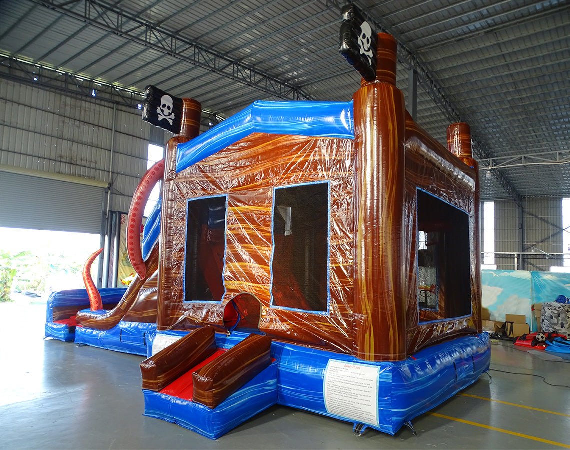 Rear view of Tentacle Typhoon Dual Lane Commercial Inflatable Combo set up in a warehouse. The inflatable displays a brown ship-like structure with blue trim, red octopus tentacles, and a bounce house area with mesh windows. Pirate flags are visible on top. Various other inflatable products and warehouse equipment can be seen in the background, demonstrating its commercial setting.