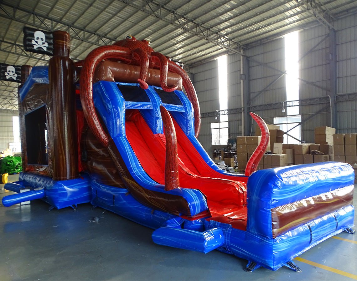 Close-up front view of Tentacle Typhoon Dual Lane Commercial Inflatable Combo (Wet/Dry) showing the entrance area and splash pool. Features a brown ship-like structure with blue trim, large red octopus tentacles wrapping around the sides, and dual slides in red and blue. A blue inflatable splash pool surrounds the base. Black pirate flags with white skull and crossbones are visible on top. Set in a warehouse with concrete flooring and various other inflatable products and equipment in the background.