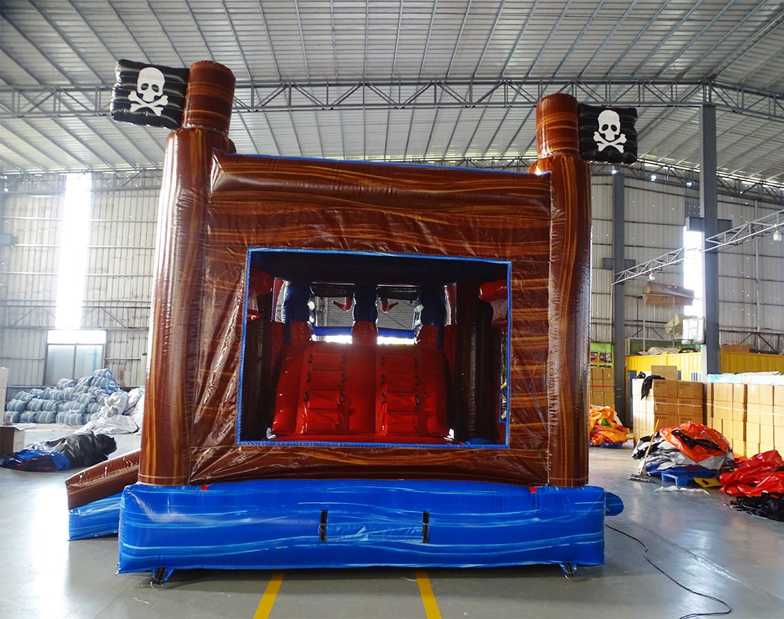 Rear view of Tentacle Typhoon Dual Lane Commercial Inflatable Combo (Wet/Dry) featuring a pirate ship theme with large octopus tentacles. The structure has dual slides, one red and one blue, emerging from a brown ship-like base. Red inflatable octopus tentacles surround the sides and front. Black pirate flags with white skull and crossbones adorn the top. Set up in a large indoor warehouse with metal roofing, bright lighting, and various other inflatable products visible in the background.