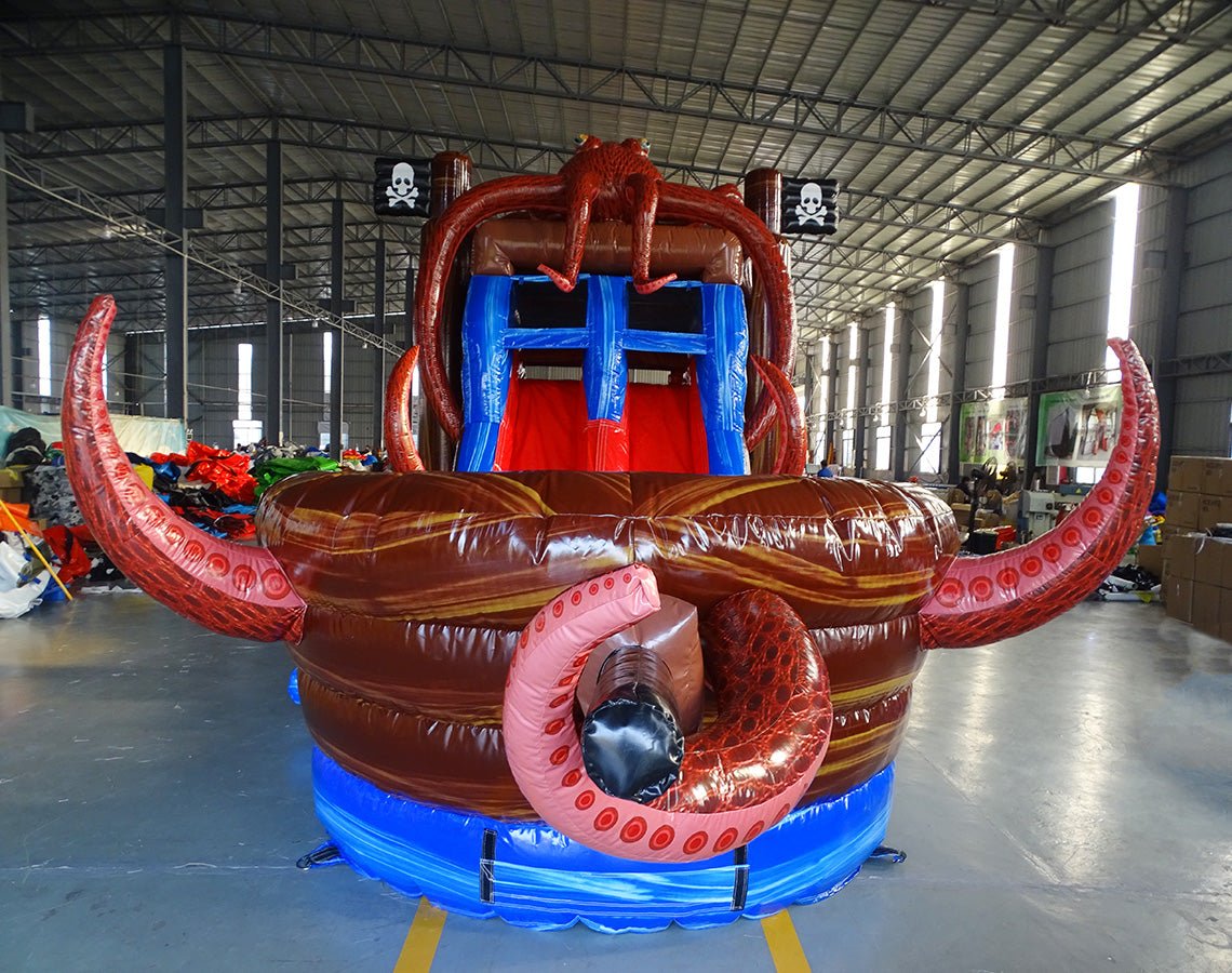 Close-up front view of Tentacle Typhoon Dual Lane Commercial Inflatable Combo showing the entrance area. Features a brown ship-like structure with blue trim, red octopus tentacles, and dual slides in red and blue. A large inflatable octopus head is visible at the top. Set in a warehouse with concrete flooring and various other inflatable products visible in the background.