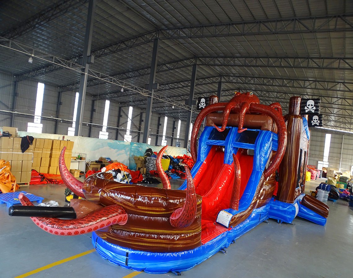 Front view of Tentacle Typhoon Dual Lane Commercial Inflatable Combo featuring a pirate ship theme with large octopus tentacles. The structure has dual slides, one red and one blue, emerging from a brown ship-like base. Inflatable octopus tentacles in red surround the sides. Pirate flags with skull and crossbones adorn the top. Set up in a large indoor warehouse with metal roofing and bright lighting visible.