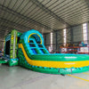 Side view of Tiki Shot Curved Dual Lane Commercial Inflatable Combo (Wet/Dry) set up in a large indoor warehouse. Features a tall tiki-faced tower with dual curved slides, a large splash pool, and vibrant tropical colors. Metal roofing and other warehouse equipment visible in the background.