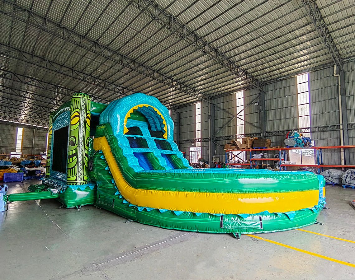 Side view of Tiki Shot Curved Dual Lane Commercial Inflatable Combo (Wet/Dry) set up in a large indoor warehouse. Features a tall tiki-faced tower with dual curved slides, a large splash pool, and vibrant tropical colors. Metal roofing and other warehouse equipment visible in the background.