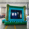 Wide-angle view of Tiki Shot Curved Dual Lane Commercial Inflatable Combo (Wet/Dry) in a spacious warehouse. Features a large tiki-faced tower with green pillars and yellow eyes, dual curved blue slides, and a circular splash pool with yellow and green accents. The inflatable structure has vibrant turquoise, green, and yellow colors. Warehouse interior visible with metal roofing, shelving units, and other equipment, providing scale and context for the inflatable's size.