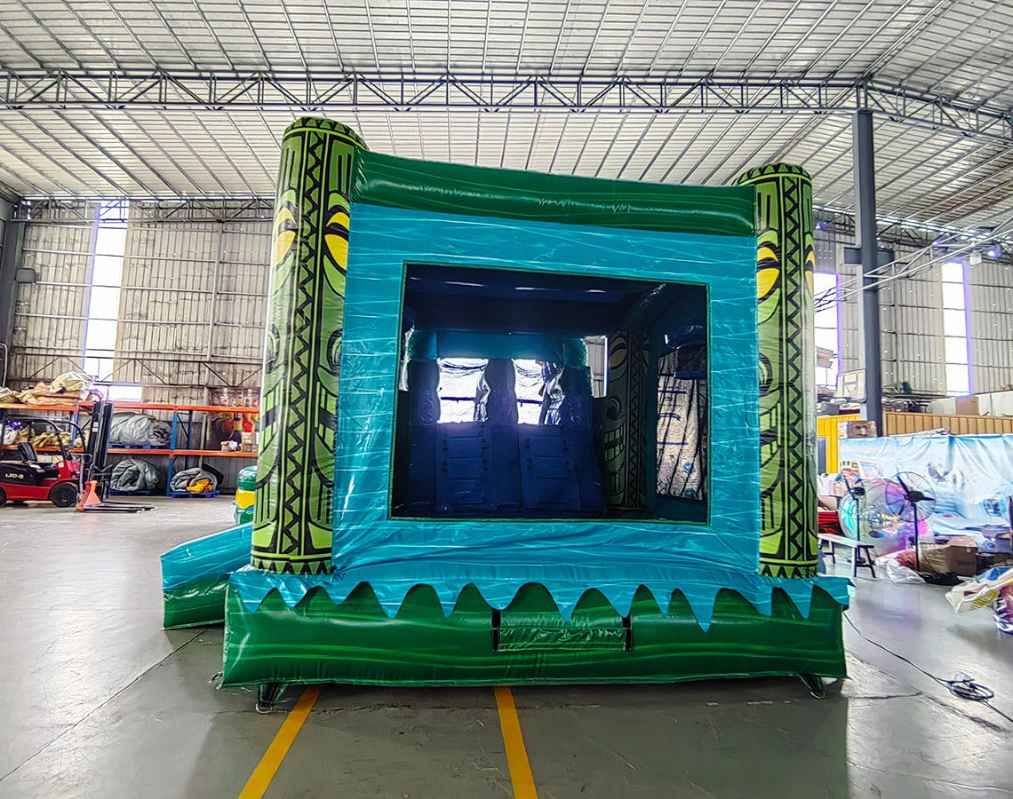 Wide-angle view of Tiki Shot Curved Dual Lane Commercial Inflatable Combo (Wet/Dry) in a spacious warehouse. Features a large tiki-faced tower with green pillars and yellow eyes, dual curved blue slides, and a circular splash pool with yellow and green accents. The inflatable structure has vibrant turquoise, green, and yellow colors. Warehouse interior visible with metal roofing, shelving units, and other equipment, providing scale and context for the inflatable's size.