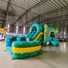 Front angled view of Tiki Shot Curved Dual Lane Commercial Inflatable Combo (Wet/Dry) in an indoor warehouse setting. Showcases the tiki face entrance, dual curved slides, and large splash pool. Other inflatable products, shelving units, and warehouse equipment visible, demonstrating its size and commercial setting.