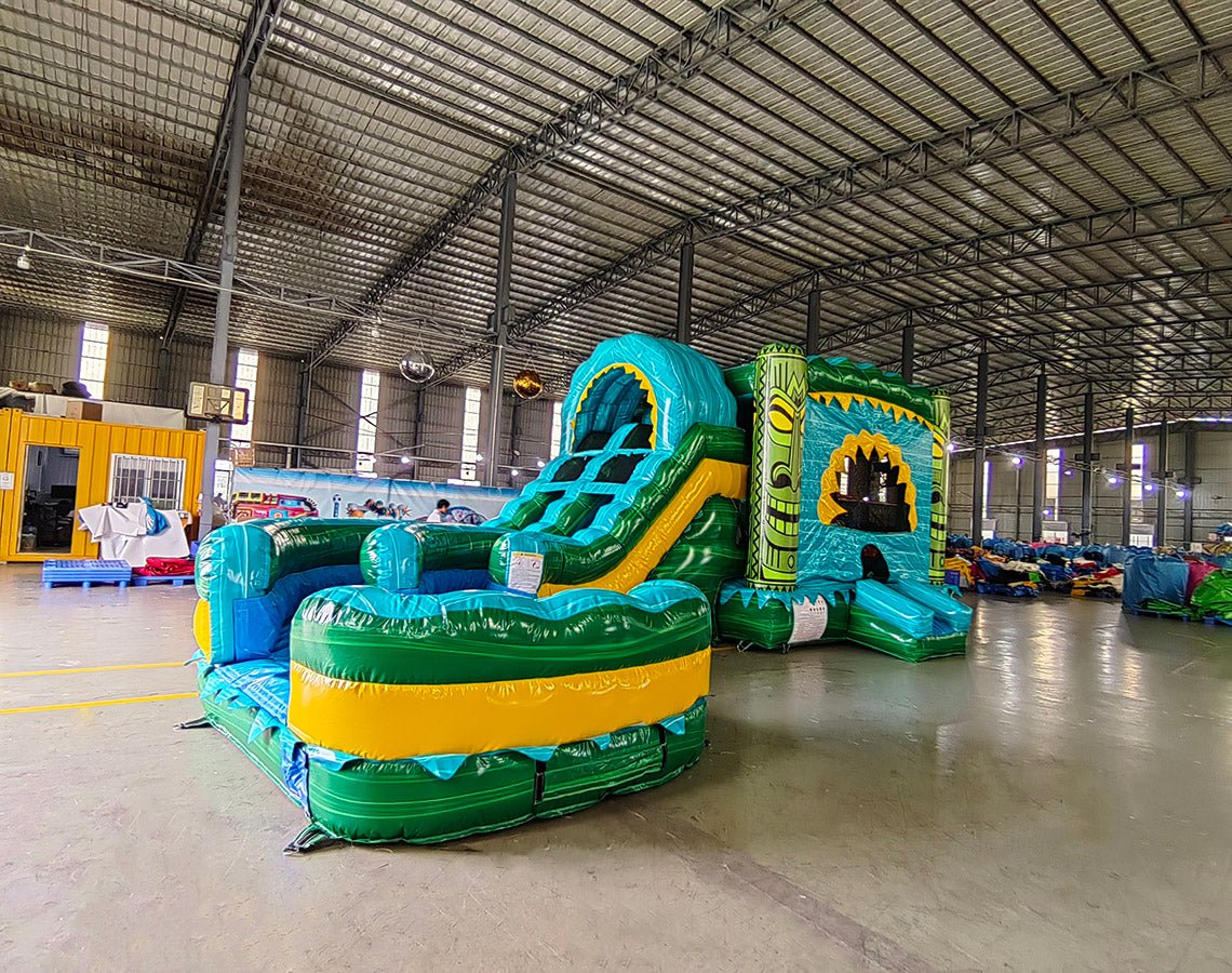 Front angled view of Tiki Shot Curved Dual Lane Commercial Inflatable Combo (Wet/Dry) in an indoor warehouse setting. Showcases the tiki face entrance, dual curved slides, and large splash pool. Other inflatable products, shelving units, and warehouse equipment visible, demonstrating its size and commercial setting.