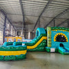 Wide-angle view of Tiki Shot Curved Dual Lane Commercial Inflatable Combo (Wet/Dry) in a spacious warehouse. Highlights the full structure including tiki face entrance, dual curved slides, splash pool, and tropical decorations. Yellow storage containers and other warehouse items visible, providing scale and context for the inflatable's size.