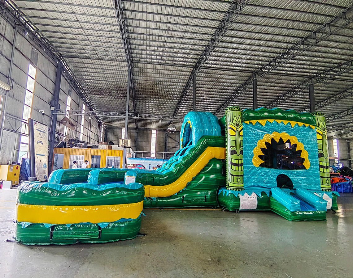 Wide-angle view of Tiki Shot Curved Dual Lane Commercial Inflatable Combo (Wet/Dry) in a spacious warehouse. Highlights the full structure including tiki face entrance, dual curved slides, splash pool, and tropical decorations. Yellow storage containers and other warehouse items visible, providing scale and context for the inflatable's size.