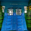 Interior view of Tiki Wave Dual Lane Commercial Inflatable Combo showing dual blue slides with safety barriers. The inflatable features a tiki theme with green tiki face designs on the sides, a blue ceiling, and a yellow wall. Set up in a warehouse with metal roofing visible above.