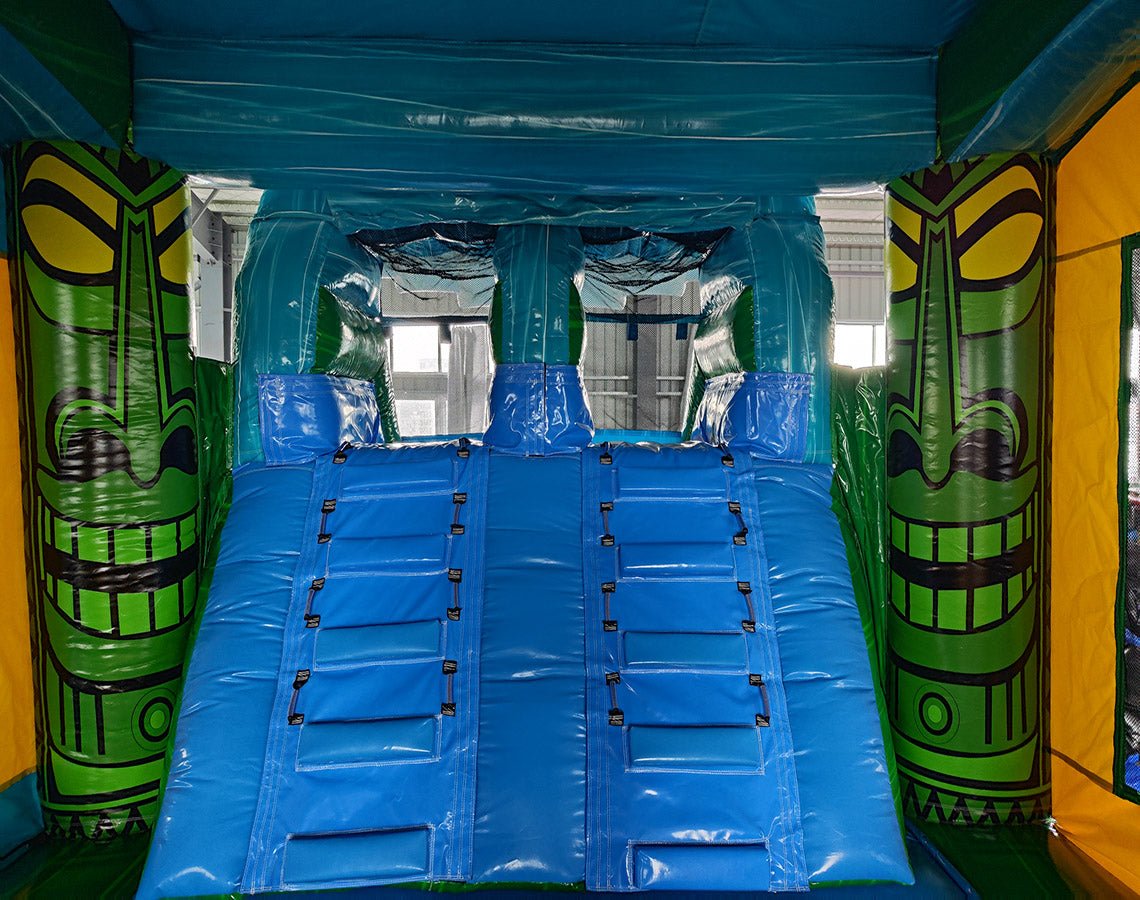 Interior view of Tiki Wave Dual Lane Commercial Inflatable Combo showing dual blue slides with safety barriers. The inflatable features a tiki theme with green tiki face designs on the sides, a blue ceiling, and a yellow wall. Set up in a warehouse with metal roofing visible above.