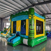 Three-quarter view of Tiki Wave Dual Lane Commercial Inflatable Combo in a spacious warehouse. The inflatable displays green tiki-style pillars with face designs, dual slides in blue and yellow, a blue wave-like top, and a yellow bounce house entrance. A large green and yellow striped splash pool area is prominent. Other inflatable products and warehouse equipment can be seen in the background.