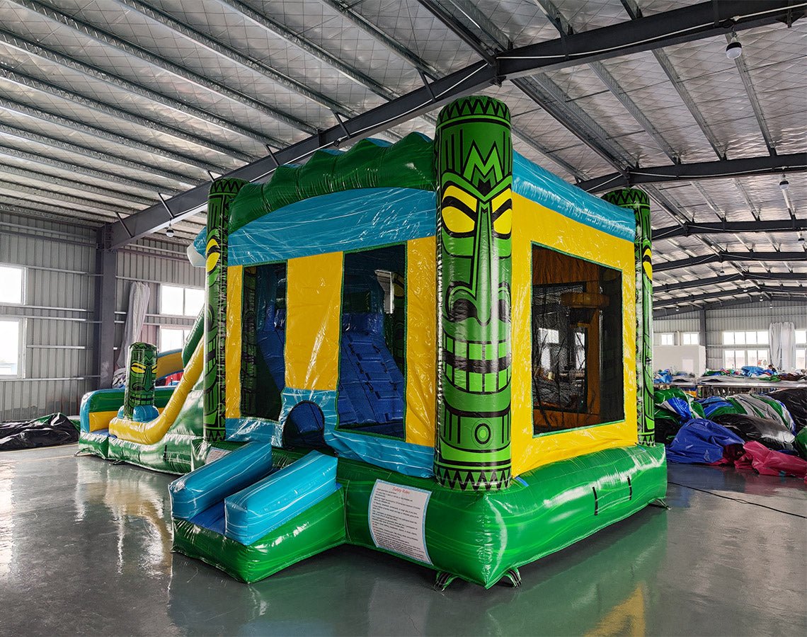 Three-quarter view of Tiki Wave Dual Lane Commercial Inflatable Combo in a spacious warehouse. The inflatable displays green tiki-style pillars with face designs, dual slides in blue and yellow, a blue wave-like top, and a yellow bounce house entrance. A large green and yellow striped splash pool area is prominent. Other inflatable products and warehouse equipment can be seen in the background.