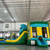 Three-quarter view of Tiki Wave Dual Lane Commercial Inflatable Combo in a spacious warehouse. The inflatable displays green tiki-style pillars, dual slides in blue and yellow, a blue wave-like top with white foam, and a yellow bounce house entrance. A large green and yellow striped splash pool area is prominent. Other inflatable products and warehouse equipment can be seen in the background.