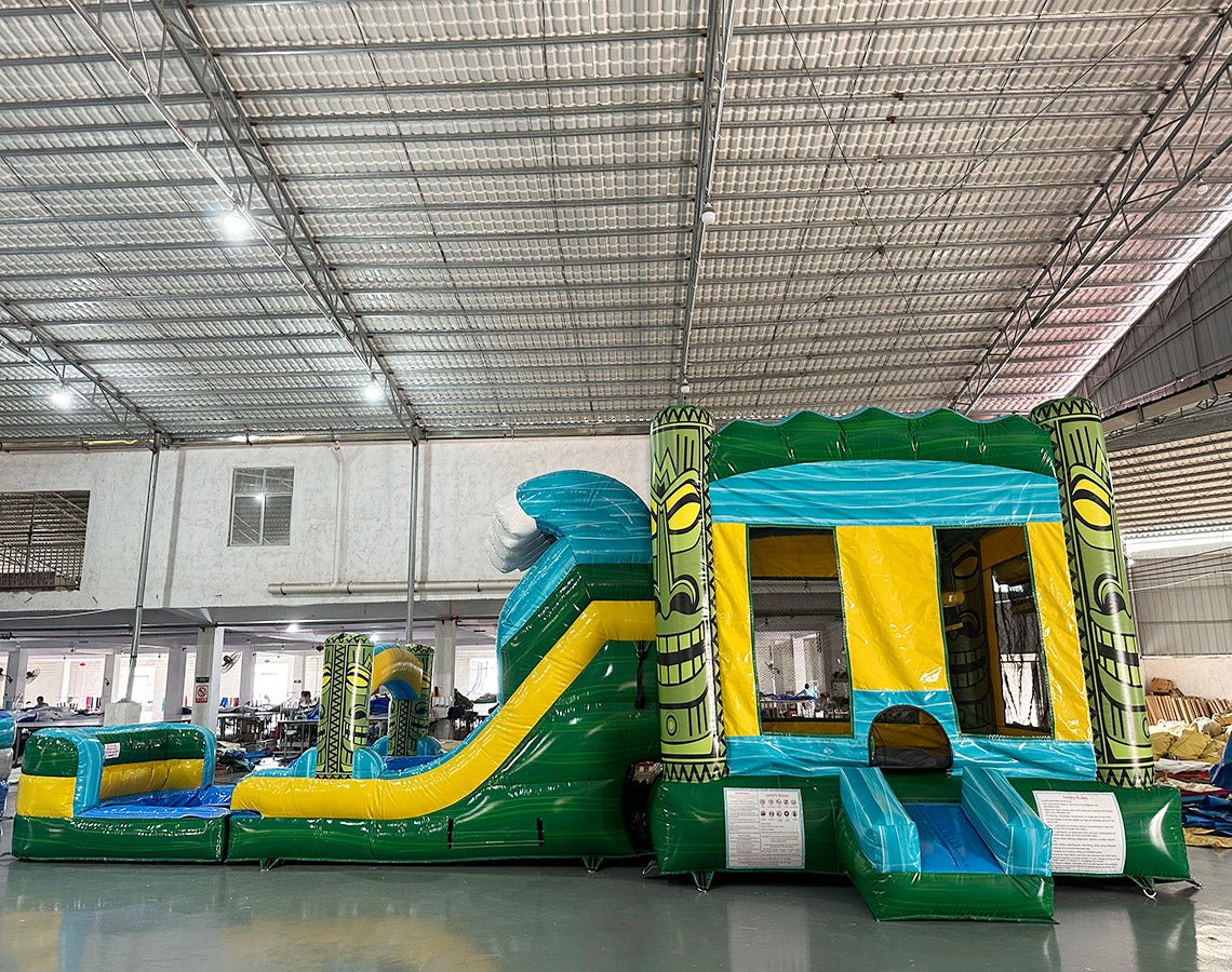 Three-quarter view of Tiki Wave Dual Lane Commercial Inflatable Combo in a spacious warehouse. The inflatable displays green tiki-style pillars, dual slides in blue and yellow, a blue wave-like top with white foam, and a yellow bounce house entrance. A large green and yellow striped splash pool area is prominent. Other inflatable products and warehouse equipment can be seen in the background.