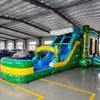 Wide-angle view of Tiki Wave Dual Lane Commercial Inflatable Combo in a warehouse setting. The structure features green tiki-style pillars, dual slides in blue and yellow, a blue wave-like top with white foam, and a large green and yellow striped splash pool. The warehouse interior with metal roofing, support beams, and various other inflatable products is visible, showcasing its commercial application.