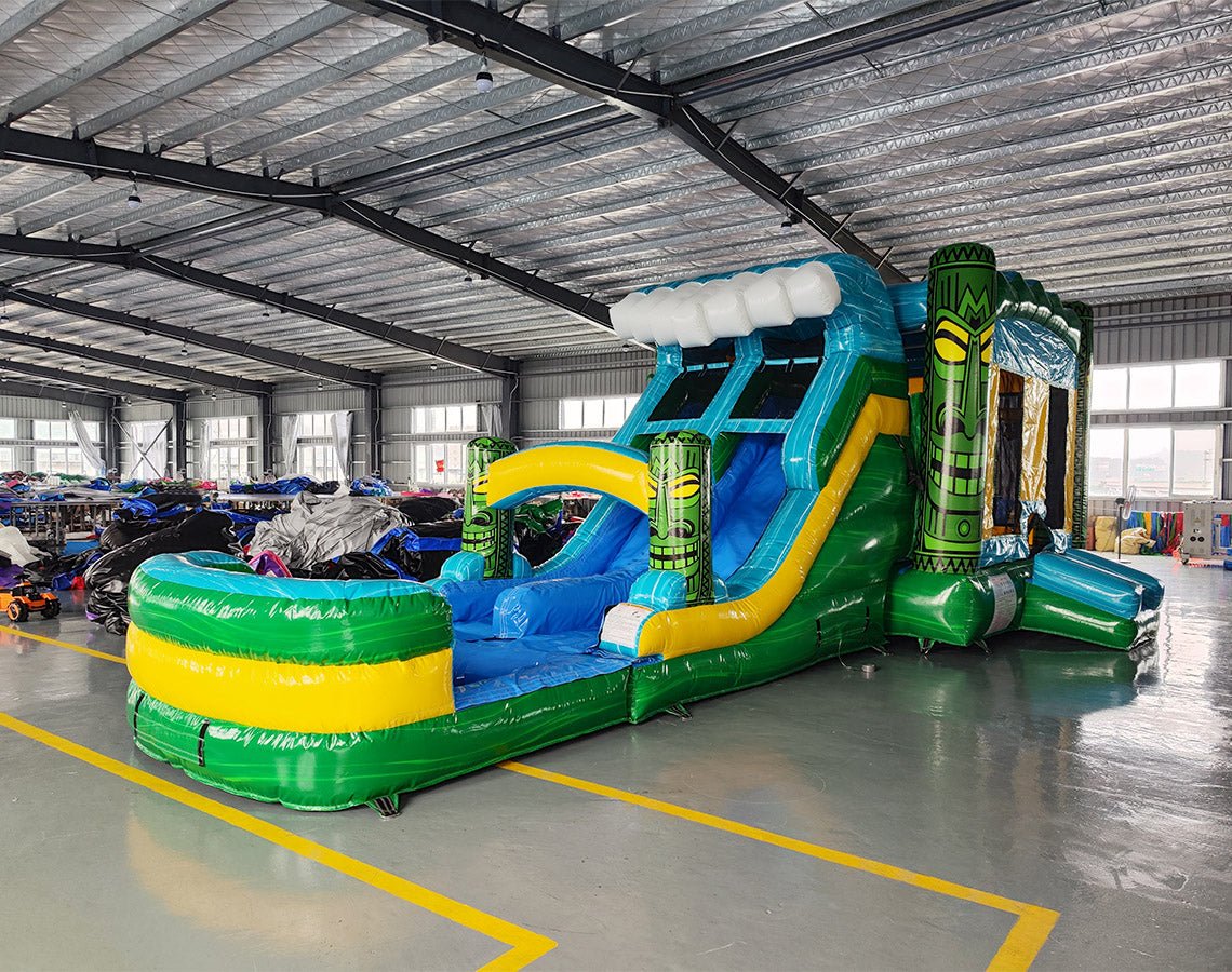 Wide-angle view of Tiki Wave Dual Lane Commercial Inflatable Combo in a warehouse setting. The structure features green tiki-style pillars, dual slides in blue and yellow, a blue wave-like top with white foam, and a large green and yellow striped splash pool. The warehouse interior with metal roofing, support beams, and various other inflatable products is visible, showcasing its commercial application.