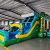 Wide-angle view of Tiki Wave Dual Lane Commercial Inflatable Combo set up in a large indoor warehouse. The inflatable features green tiki-style pillars, dual slides in blue and yellow, and a blue wave-like top with white inflatable foam. A green and yellow striped splash pool is visible at the base. Various other inflatable products and warehouse equipment are visible in the background.