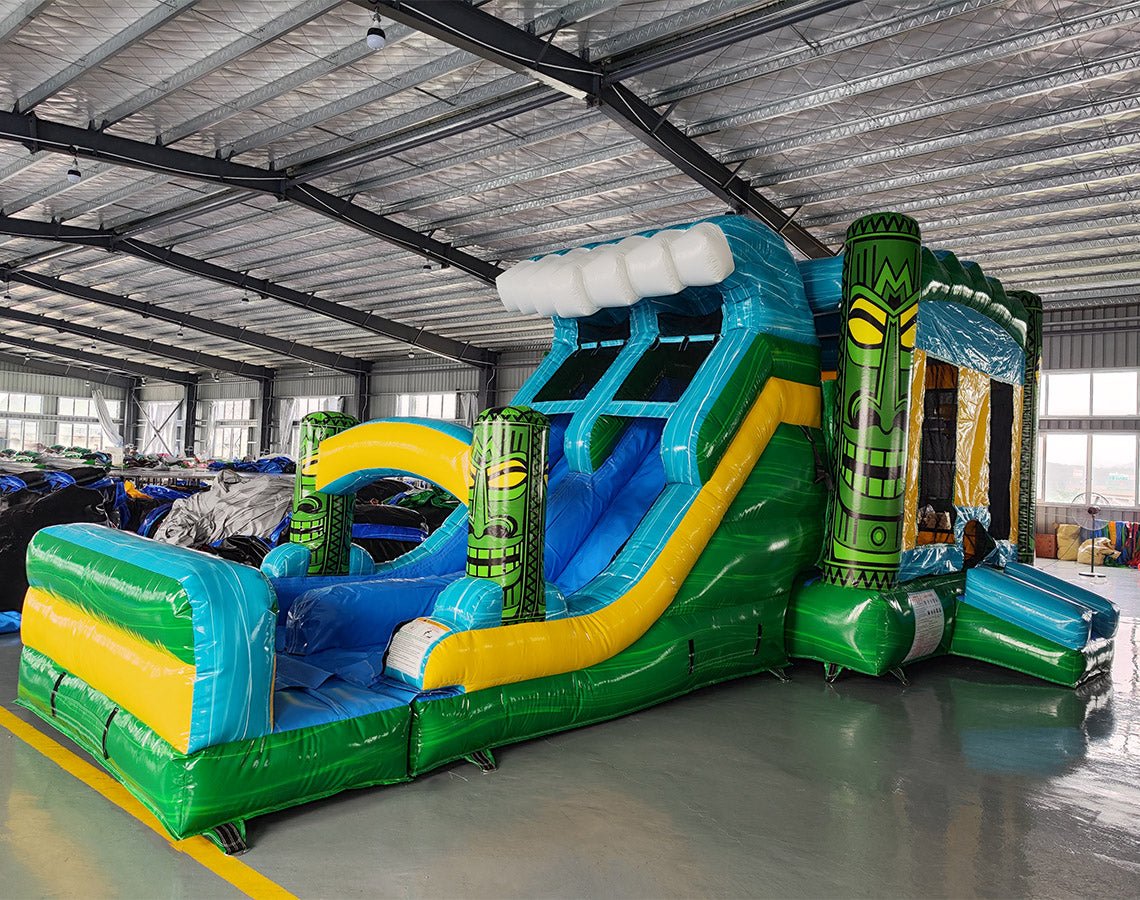 Wide-angle view of Tiki Wave Dual Lane Commercial Inflatable Combo set up in a large indoor warehouse. The inflatable features green tiki-style pillars, dual slides in blue and yellow, and a blue wave-like top with white inflatable foam. A green and yellow striped splash pool is visible at the base. Various other inflatable products and warehouse equipment are visible in the background.