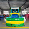 Front view of Tiki Wave Dual Lane Commercial Inflatable Combo set up in a large indoor warehouse. The inflatable features green tiki-style pillars, dual slides in blue and yellow, and a blue wave-like top with white inflatable foam. A green and yellow striped splash pool is visible at the base. Various other inflatable products and equipment are visible in the background.